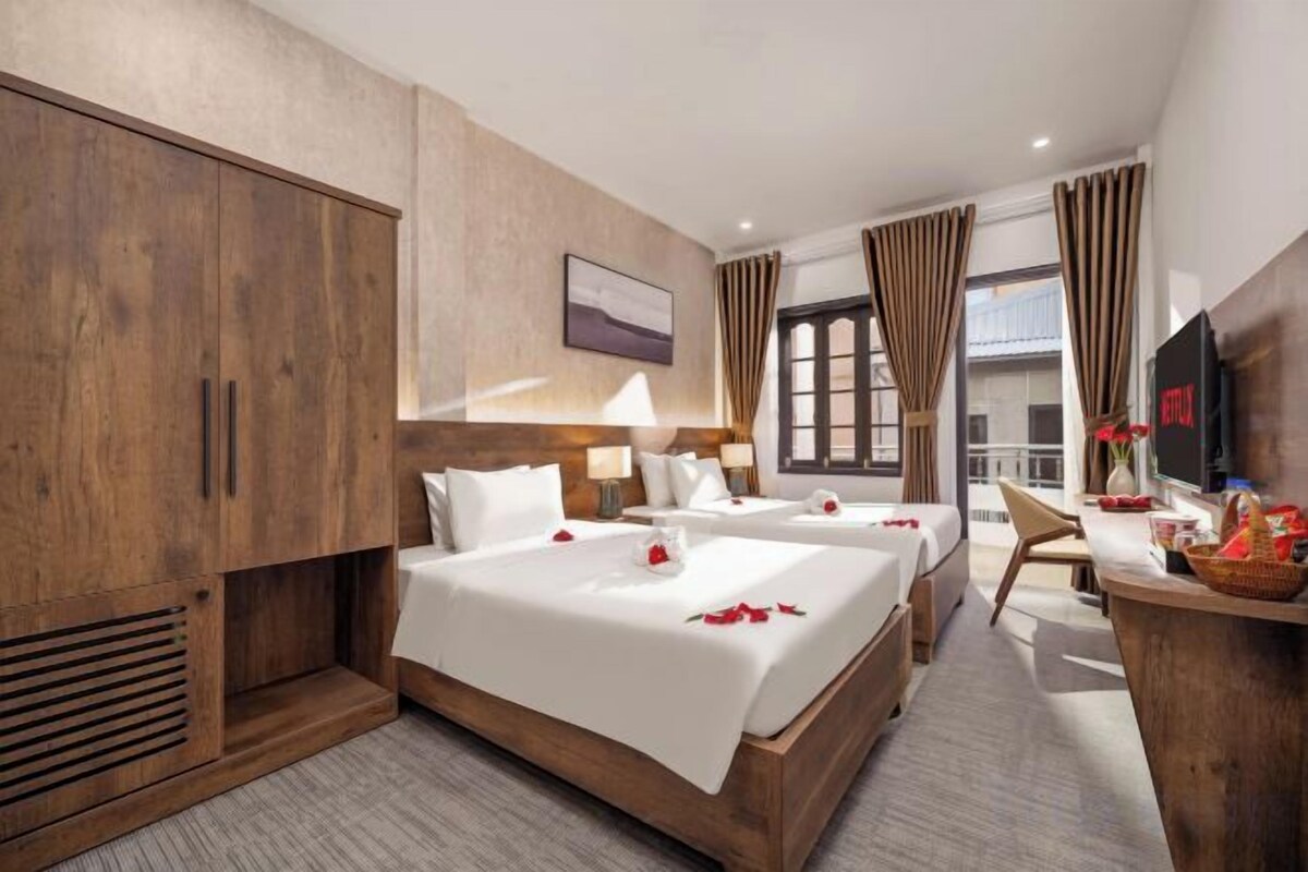 A hotel room with two beds, a wooden wardrobe, a desk with a TV, and a window with curtains.