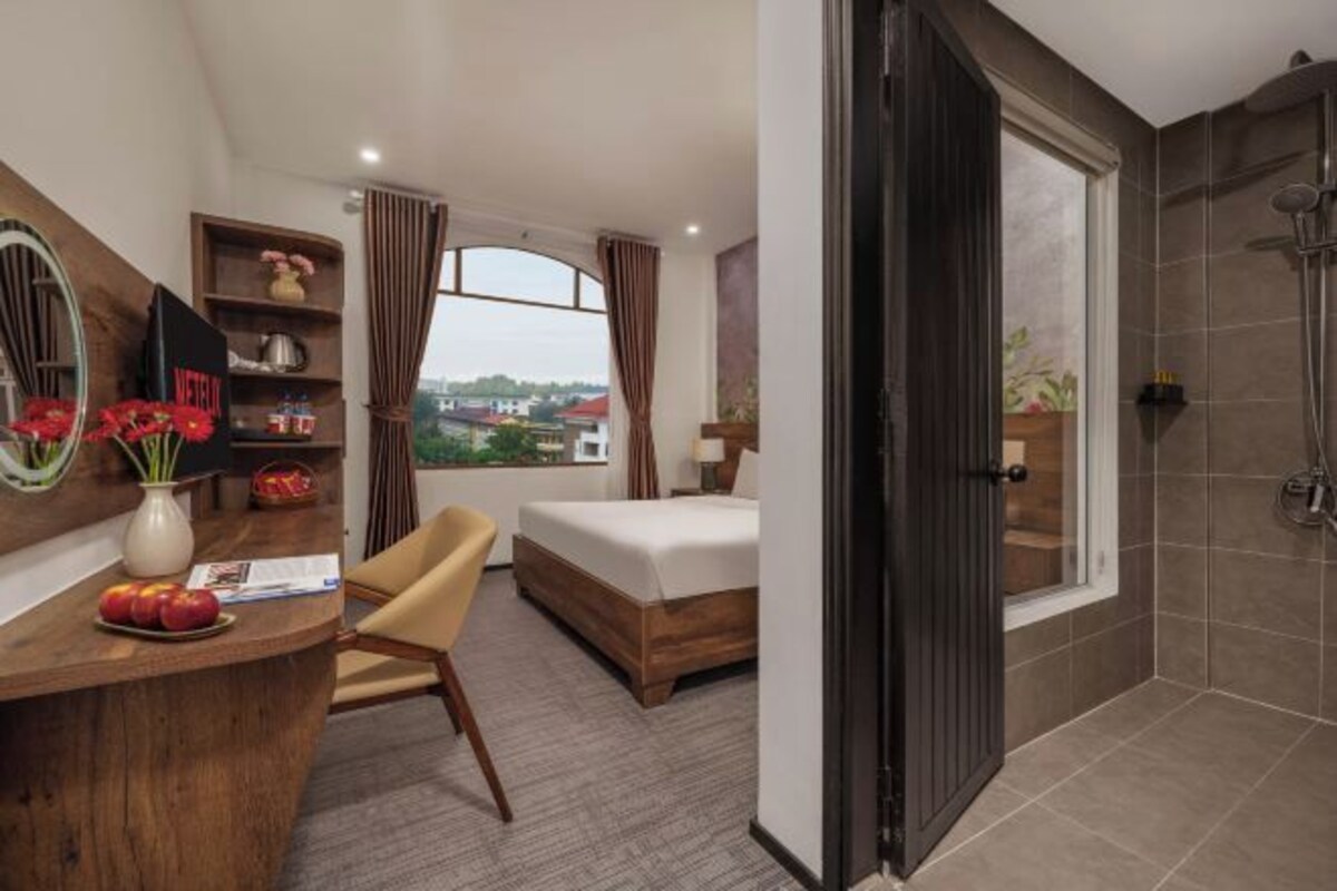 A hotel room with a bed, a desk with a vase of flowers, a chair, a window with curtains, and a shower area.