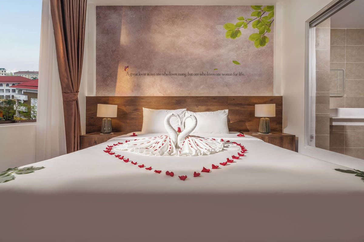 A hotel room with a large bed, two bedside lamps, a decorative wall with a quote, and a view of buildings outside.