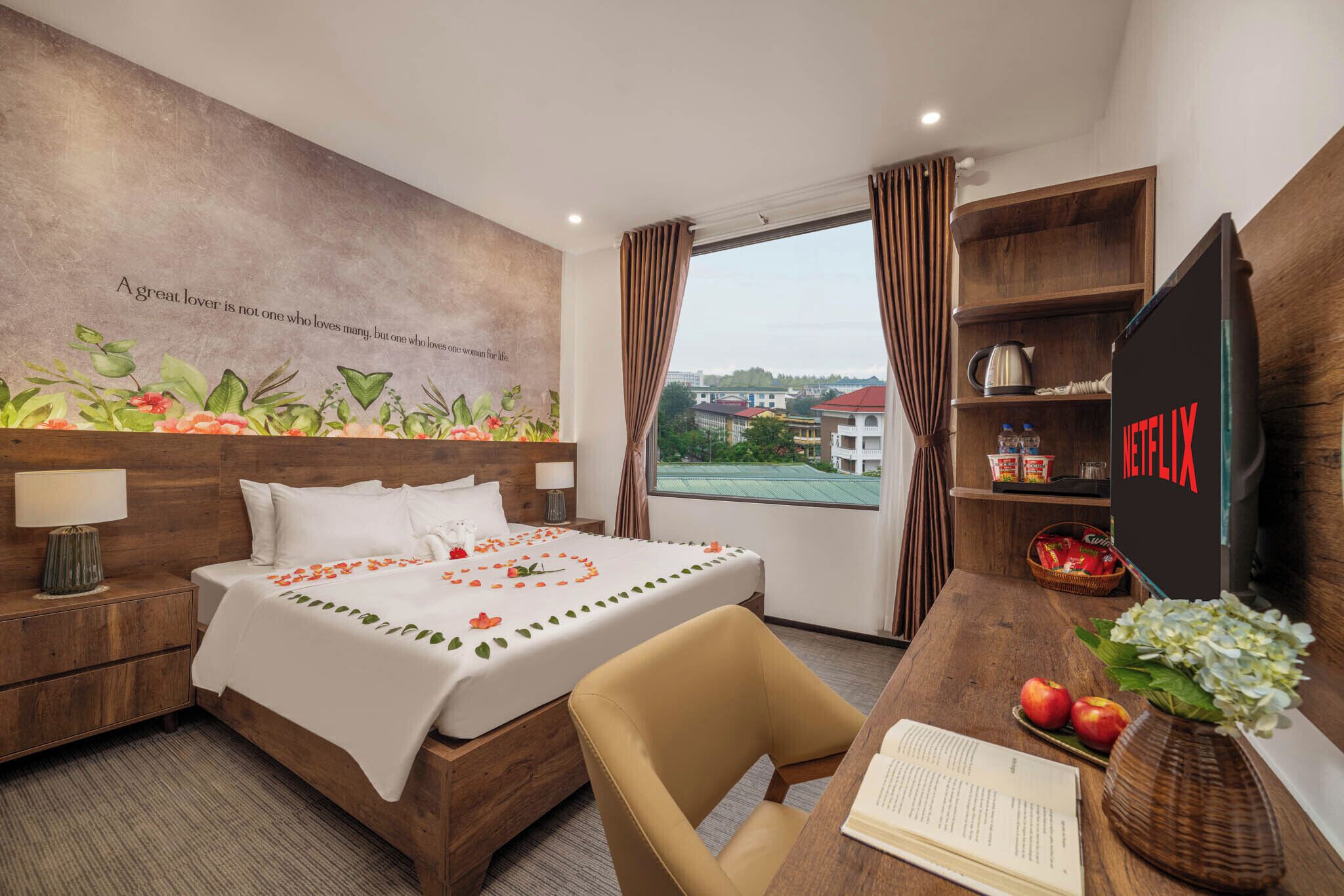 A hotel room with a bed, a desk with a laptop, a bookshelf, and a view of buildings outside.