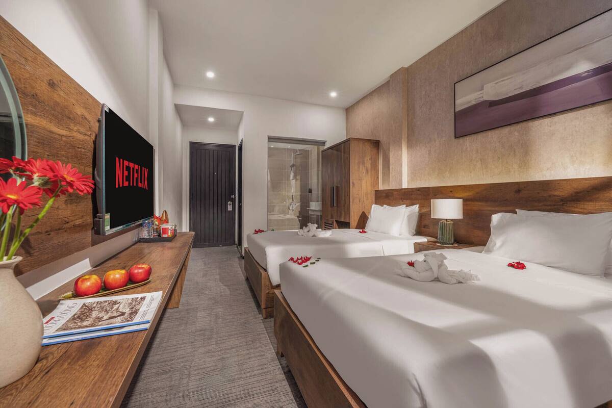 A hotel room with a wooden headboard, two beds with white linens, a flat-screen TV mounted on the wall, a wooden desk with a vase of flowers, and a door leading to a shower area.