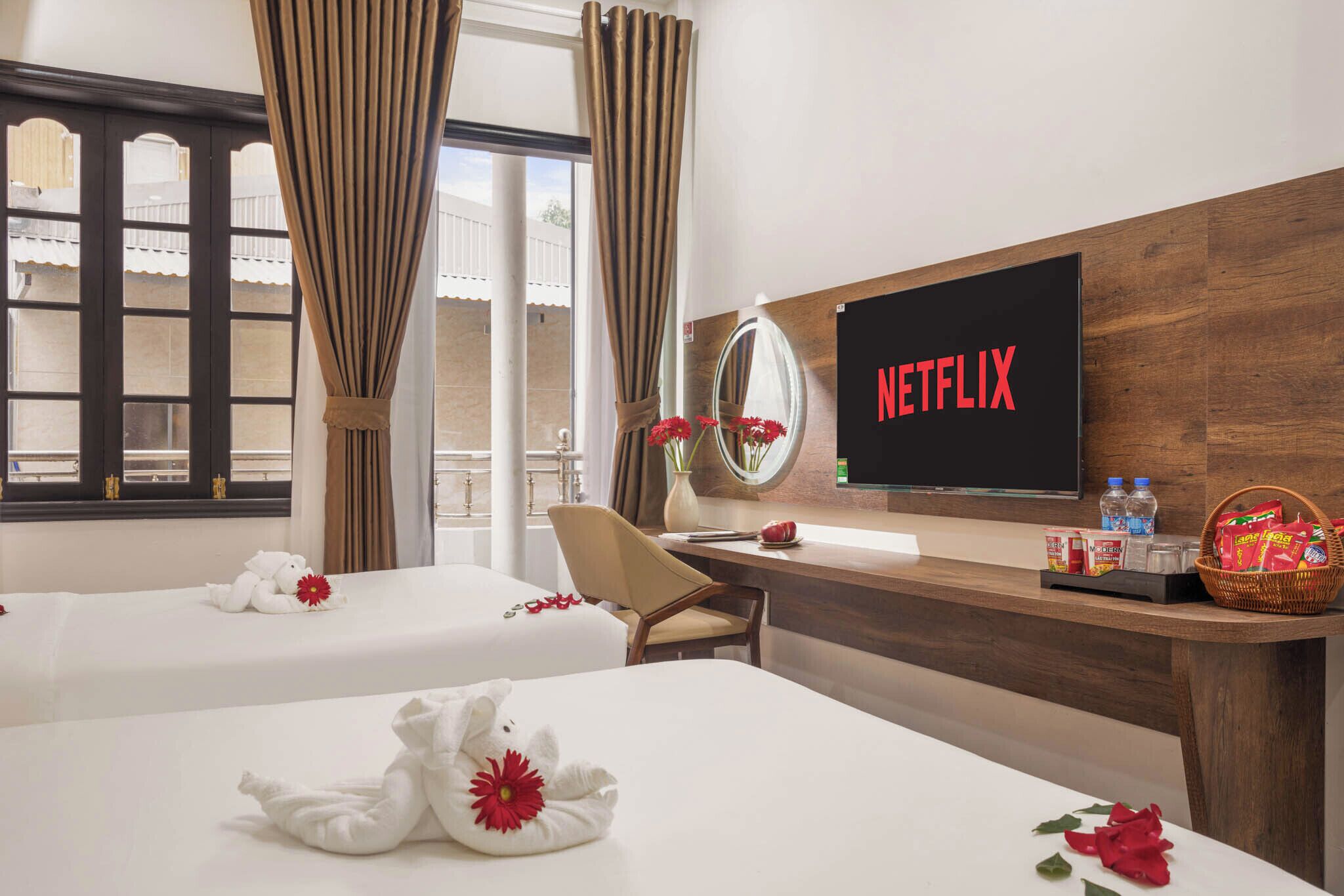 Habitació d'hotel amb dos llits, un escriptori amb una televisió que mostra 'NETFLIX', un mirall, una cadira i una finestra amb cortines.