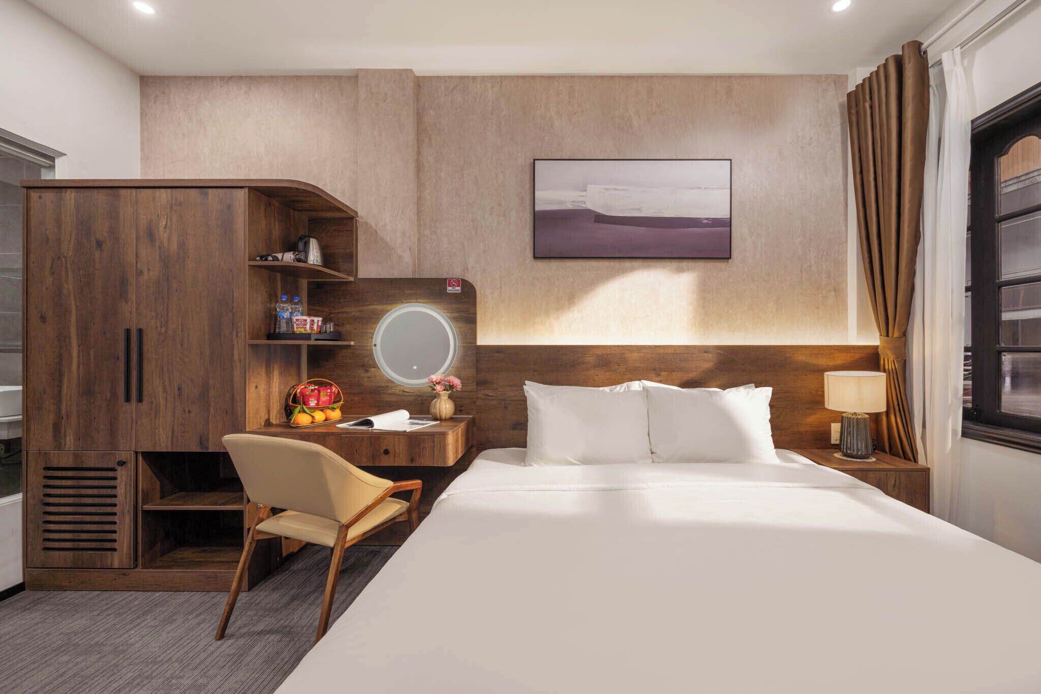 A hotel room with a bed, a desk with a chair, a wardrobe, a round mirror, a nightstand with a lamp, and a painting on the wall.