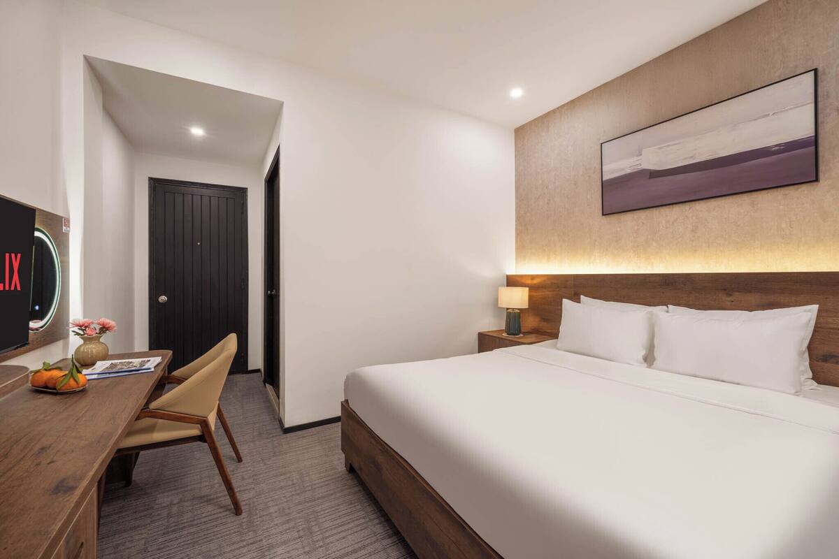 A hotel room with a bed, a desk with a chair, a TV, and a painting on the wall.