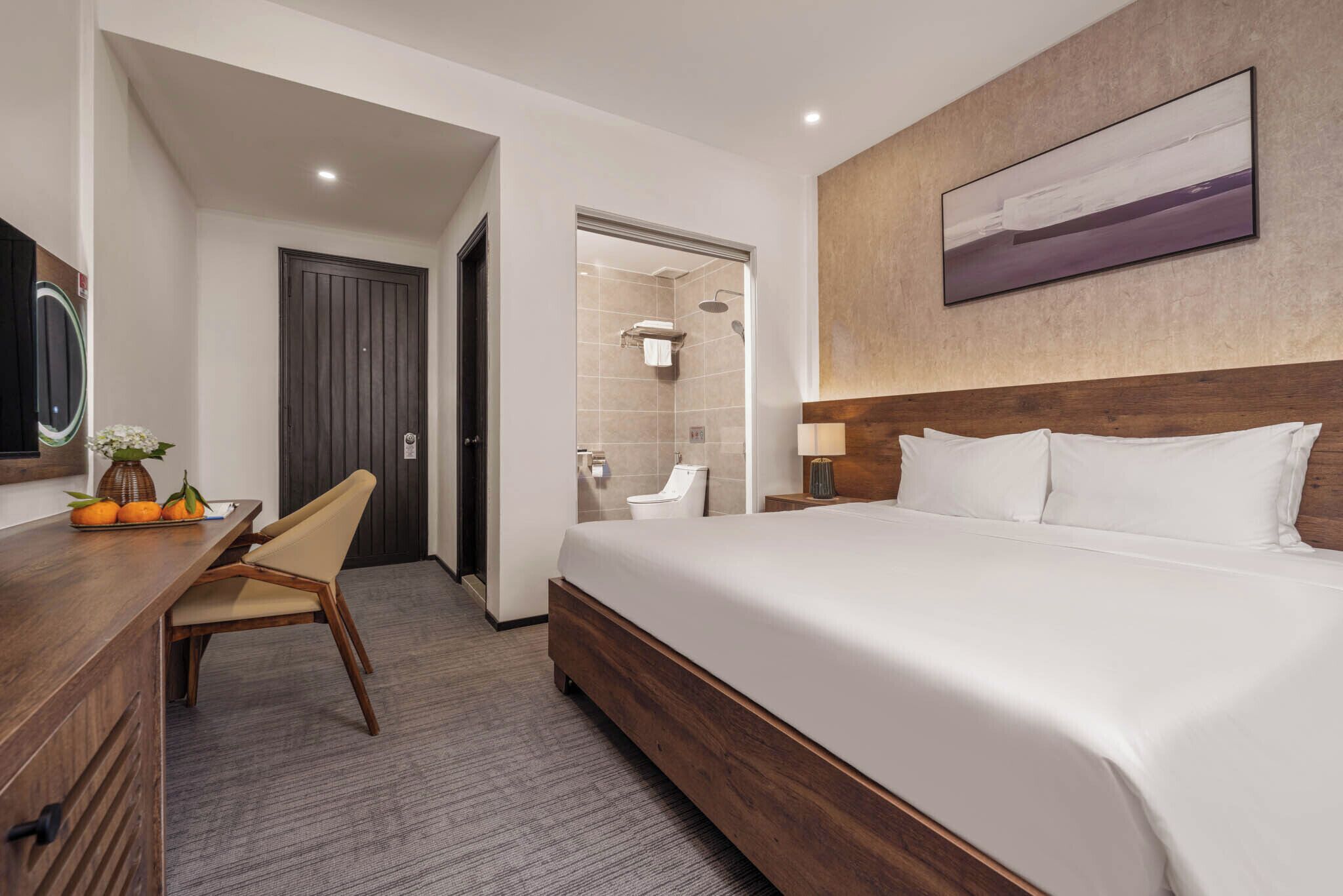 A hotel room with a bed, a desk with a chair, a bathroom with a shower, and a painting on the wall.