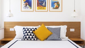 Hotellihuone, jossa on puinen sängynpääty, valkoisilla lakanoiden ja kuviollisilla tyynyillä varustettu sänky sekä kolme kehystettyä julistetta seinällä, joissa on kuvia Caprista, Napolista ja Sorrentosta.