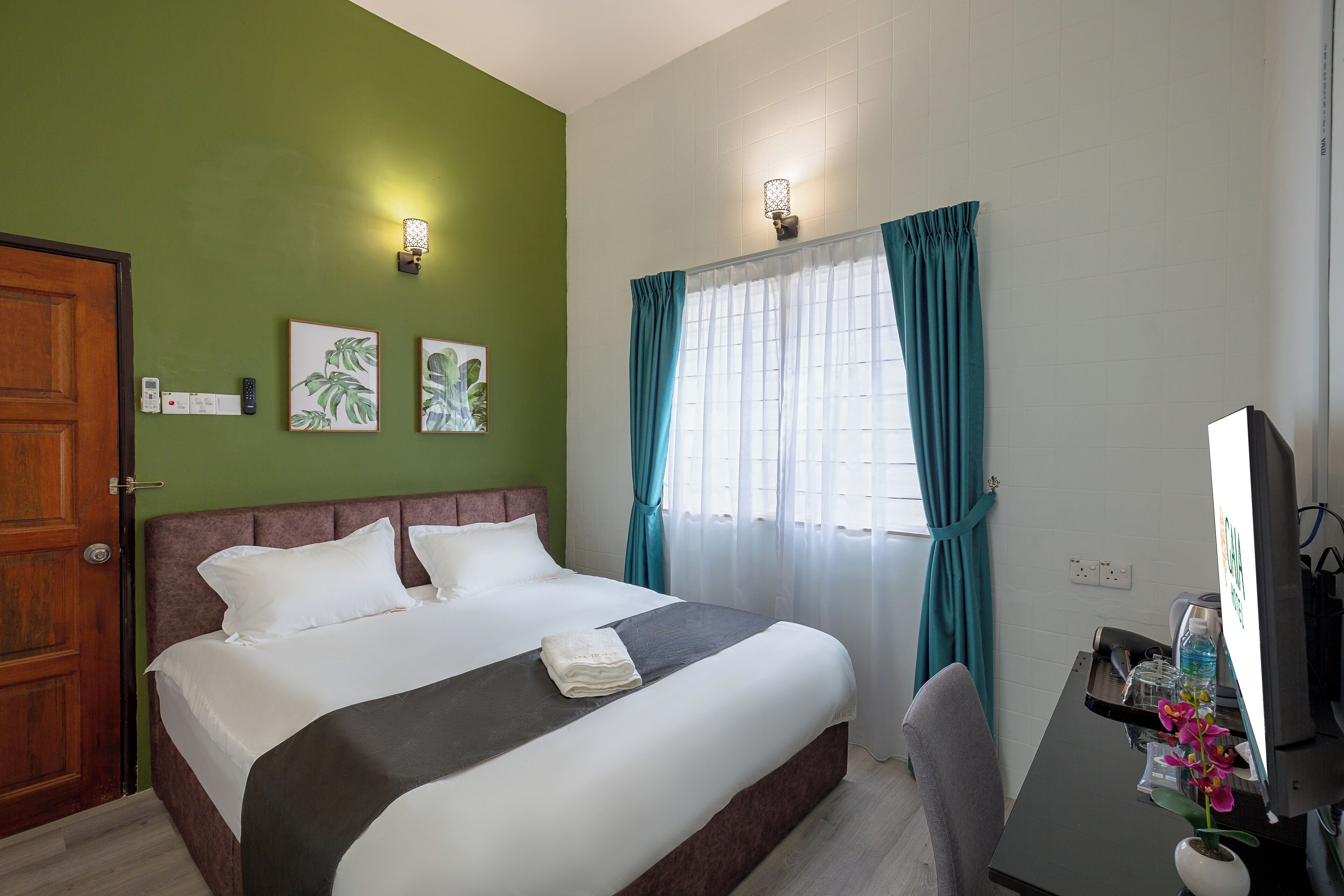 A hotel room with a bed, a desk with a computer, a chair, a window with curtains, and wall-mounted lights.