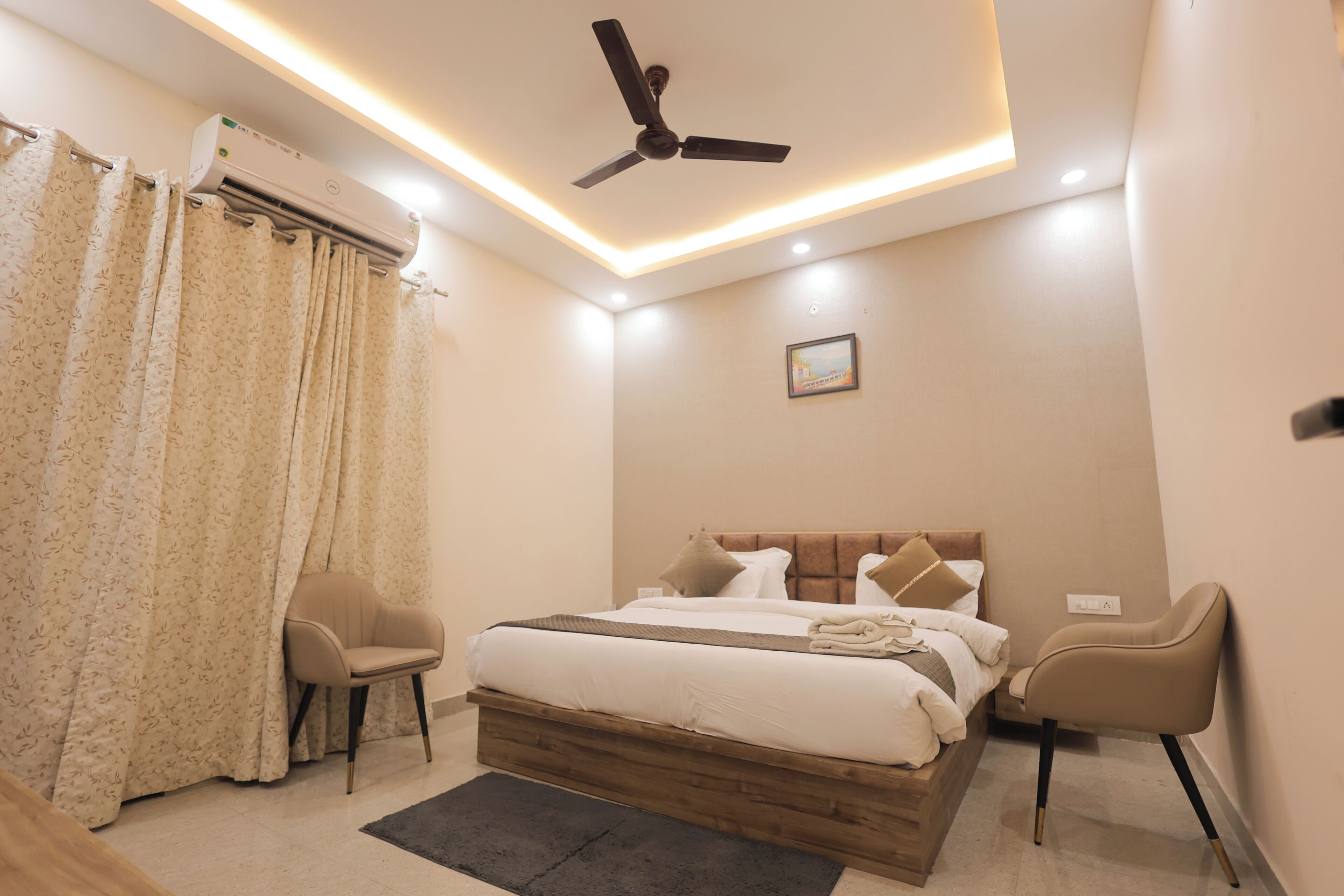 A bedroom with a bed, two chairs, a ceiling fan, an air conditioning unit, and a small picture on the wall.