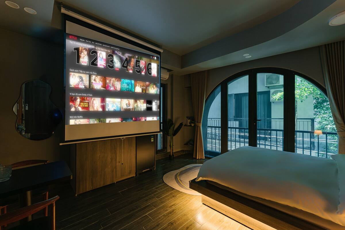 A hotel room with a large wall-mounted TV displaying a Netflix interface, a bed with a white comforter, a wooden floor, and a view of the outdoors through a large window.
