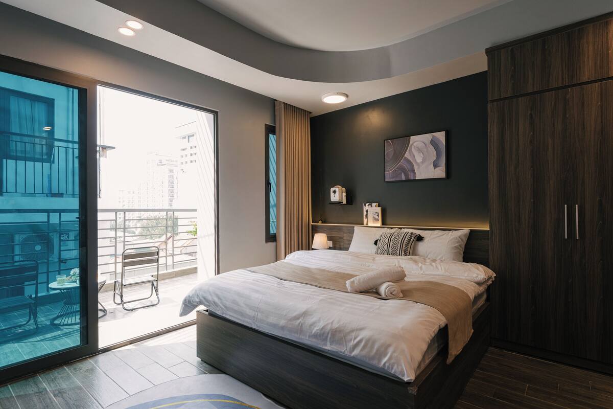 A modern hotel room with a large bed, a balcony with a view of the city, a dark wood wardrobe, and a wall-mounted shelf with a lamp.