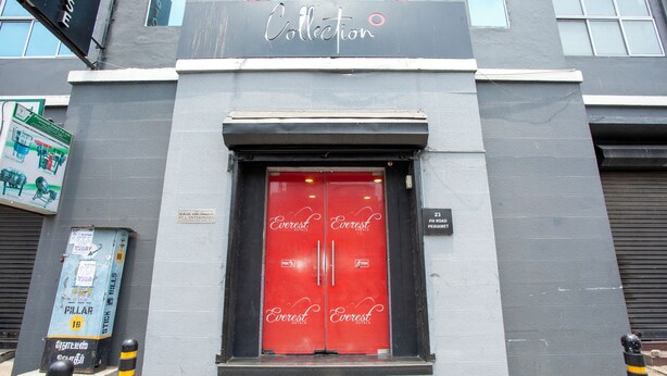 Entrada de un hotel con una puerta roja, un cartel sobre la puerta y bolardos en la parte frontal.