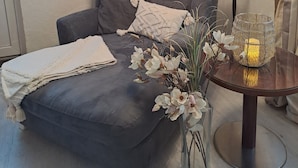 Una habitación con un sofá gris, una lámpara, un jarrón con flores y un mural en la pared.