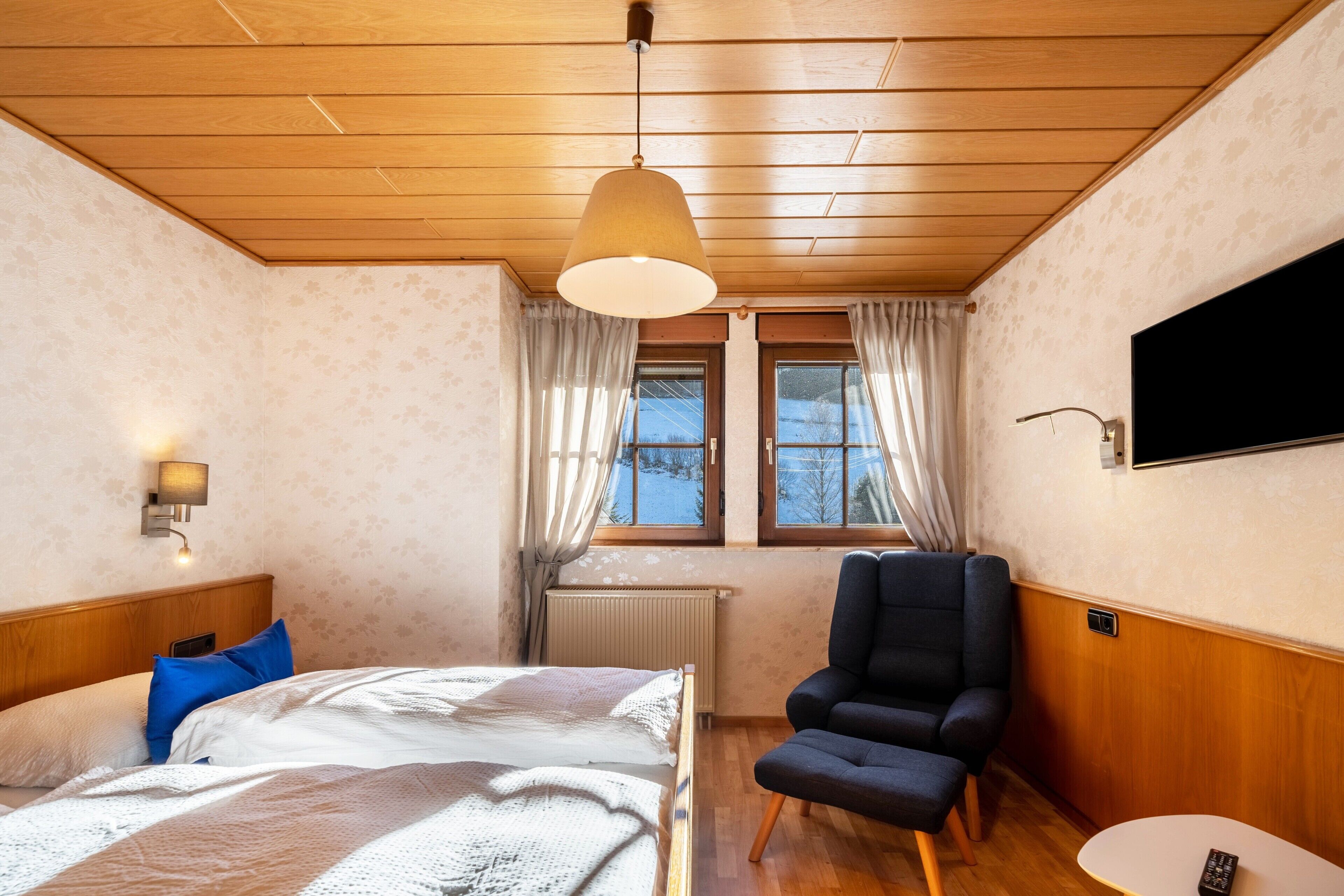 Un dormitorio con cama, una silla, un televisor y una ventana con cortinas.