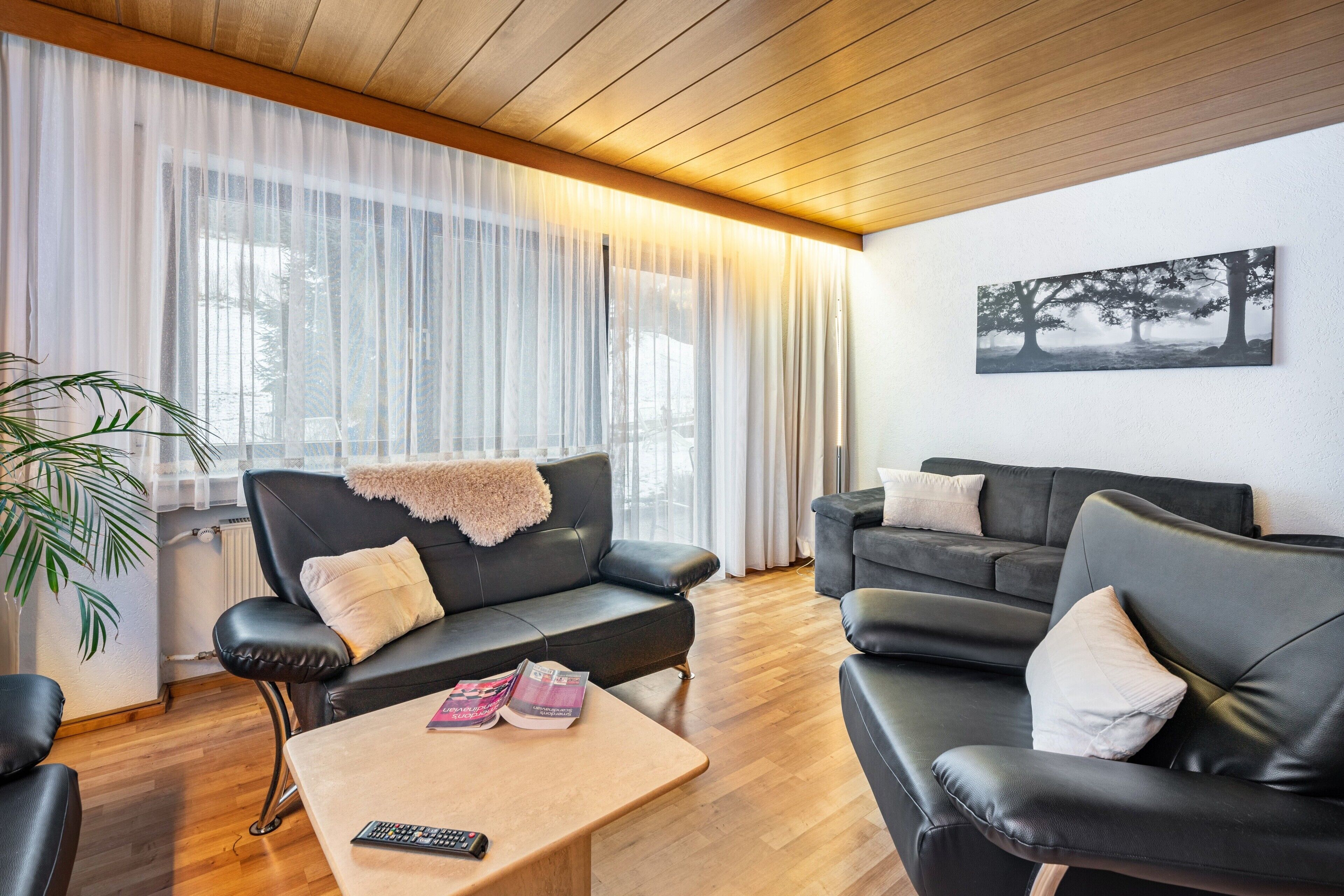 A modern living room with a wooden ceiling, black leather sofas, a coffee table with magazines, and a large window with sheer curtains.