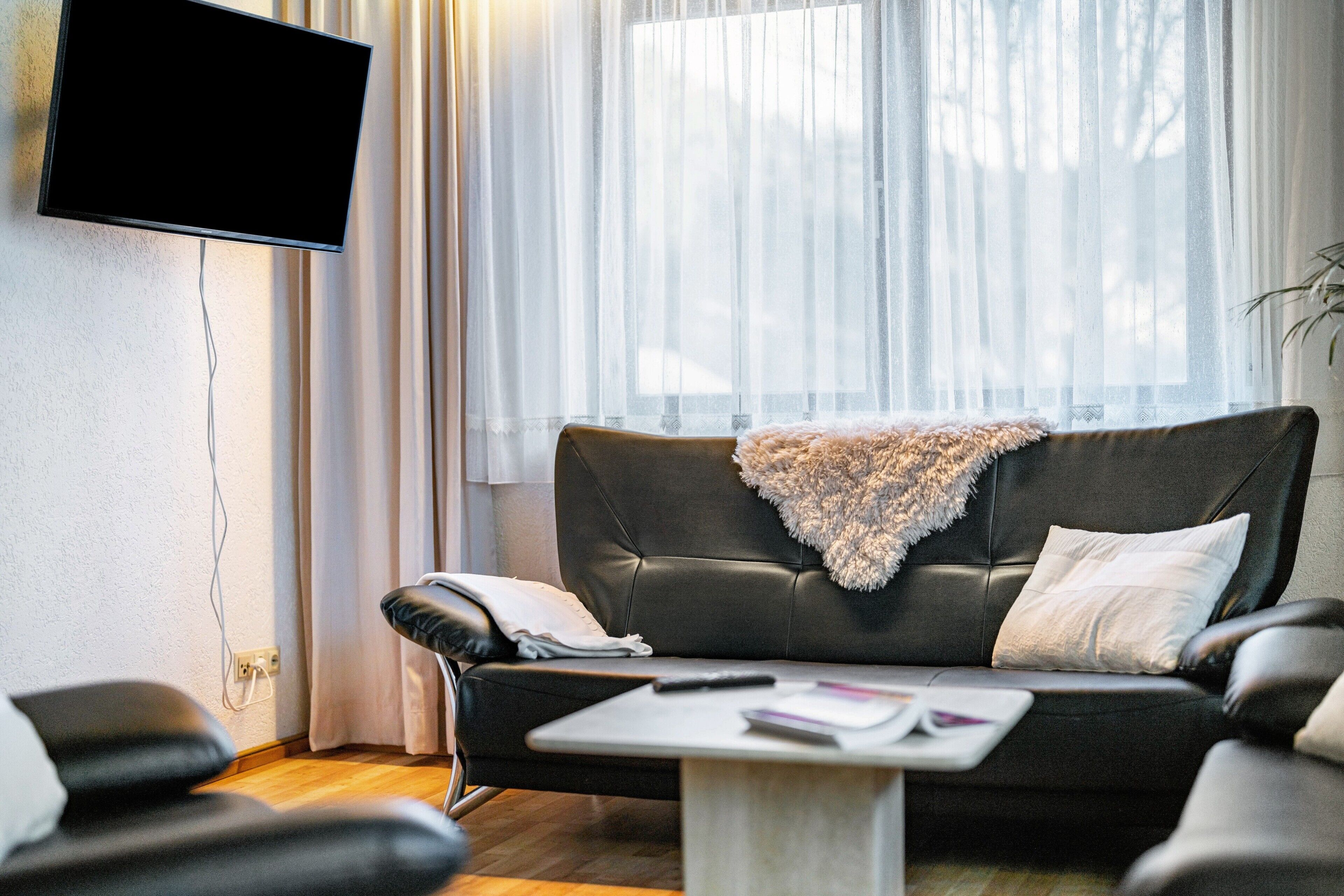 Een woonkamer met een zwarte leren bank, een witte salontafel, een wandgemonteerde flatscreen-tv en een raam met dunne gordijnen.
