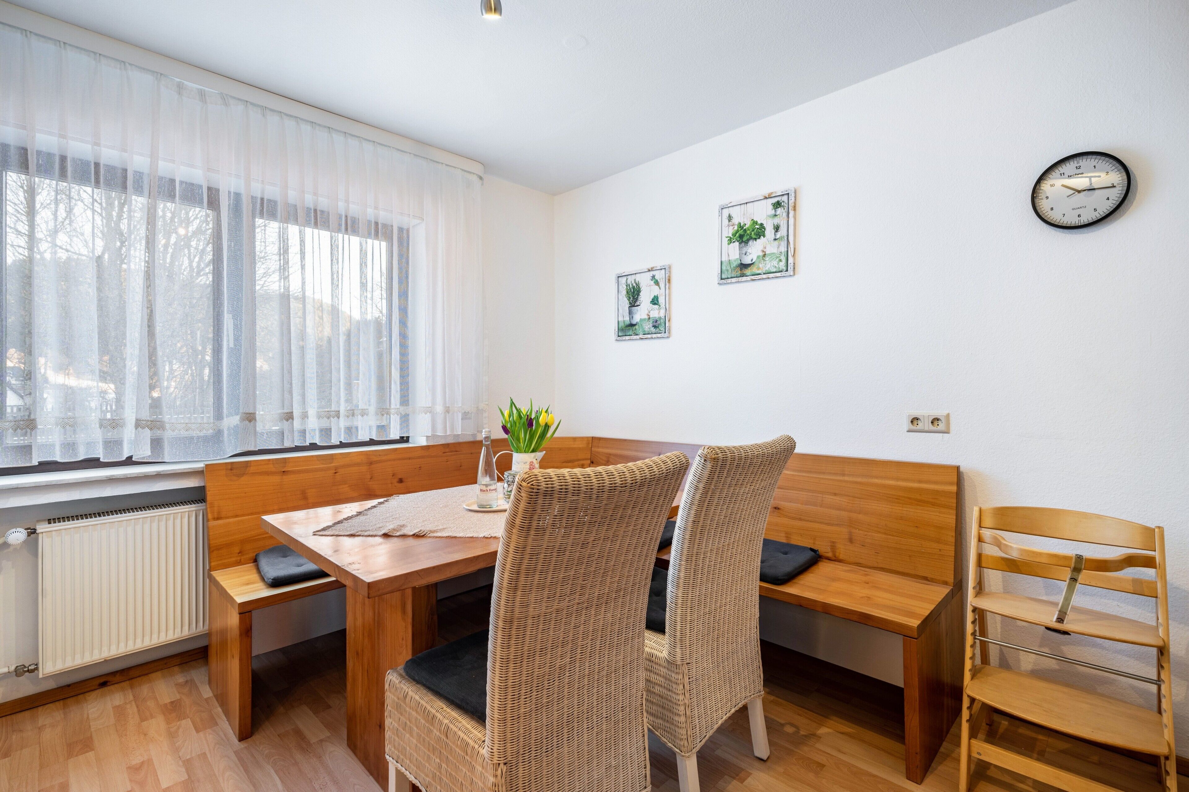 A dining area with a wooden table, wicker chairs, a bench, and a wall clock.