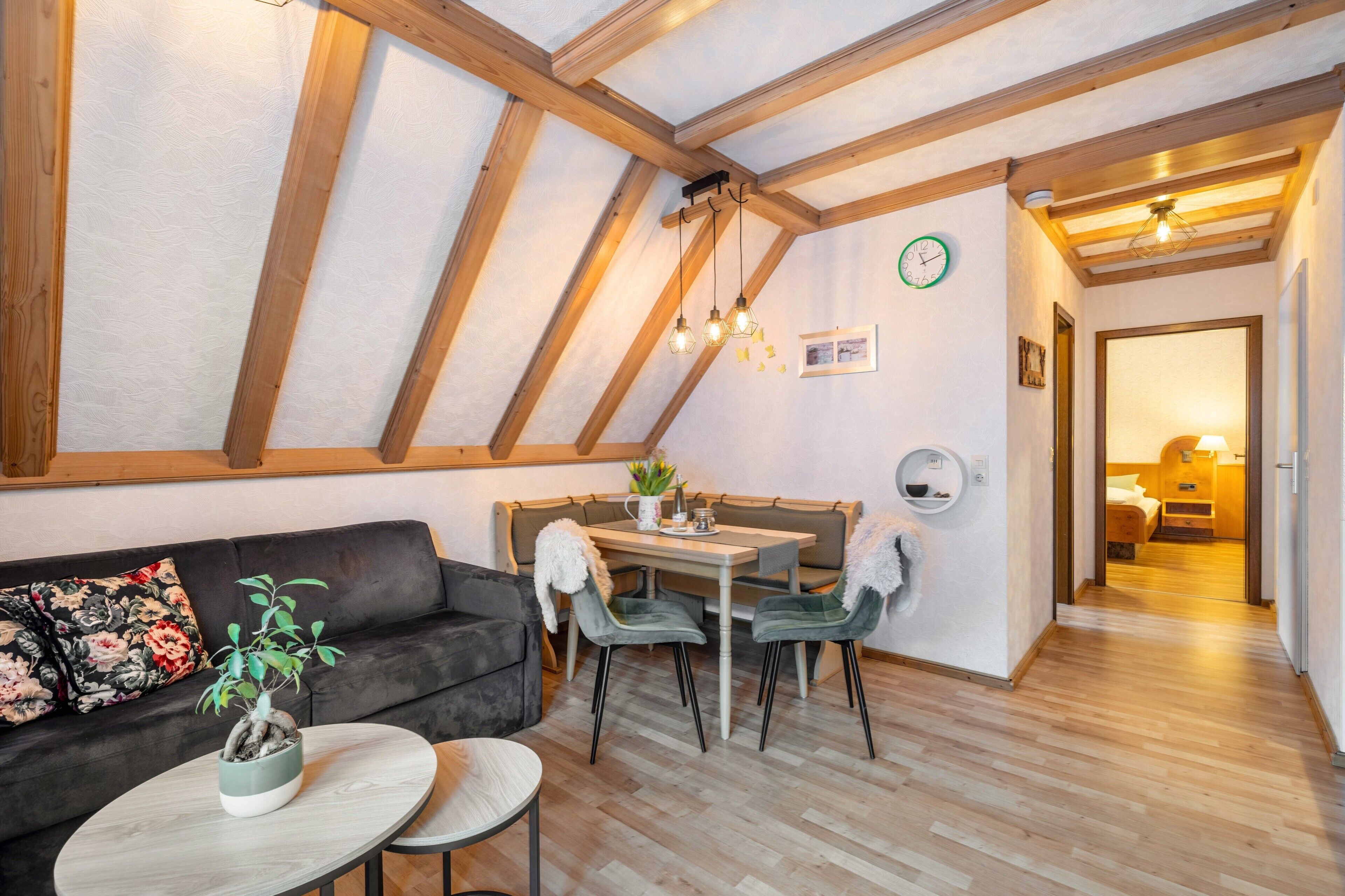 Een gezellige kamer met houten plafond, een bank, een eettafel met stoelen en een kleine ronde tafel met een plant.
