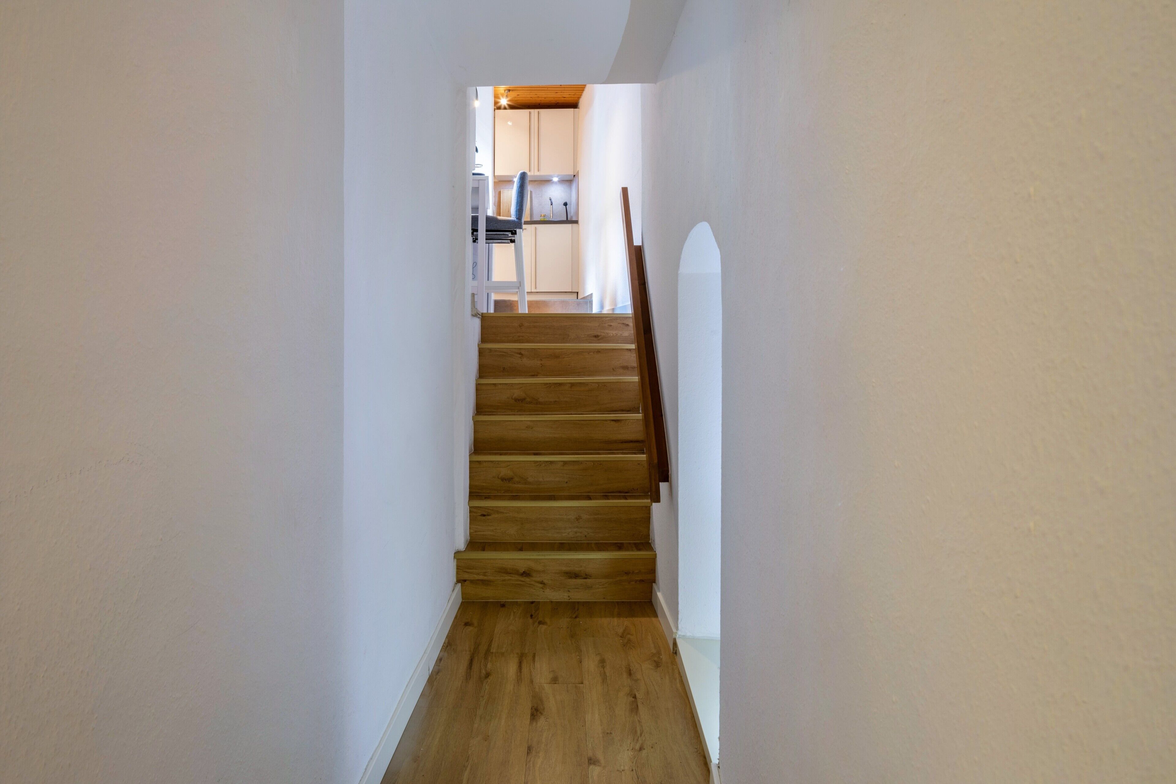 A wooden staircase leading to a kitchen area with white walls and a wooden floor.