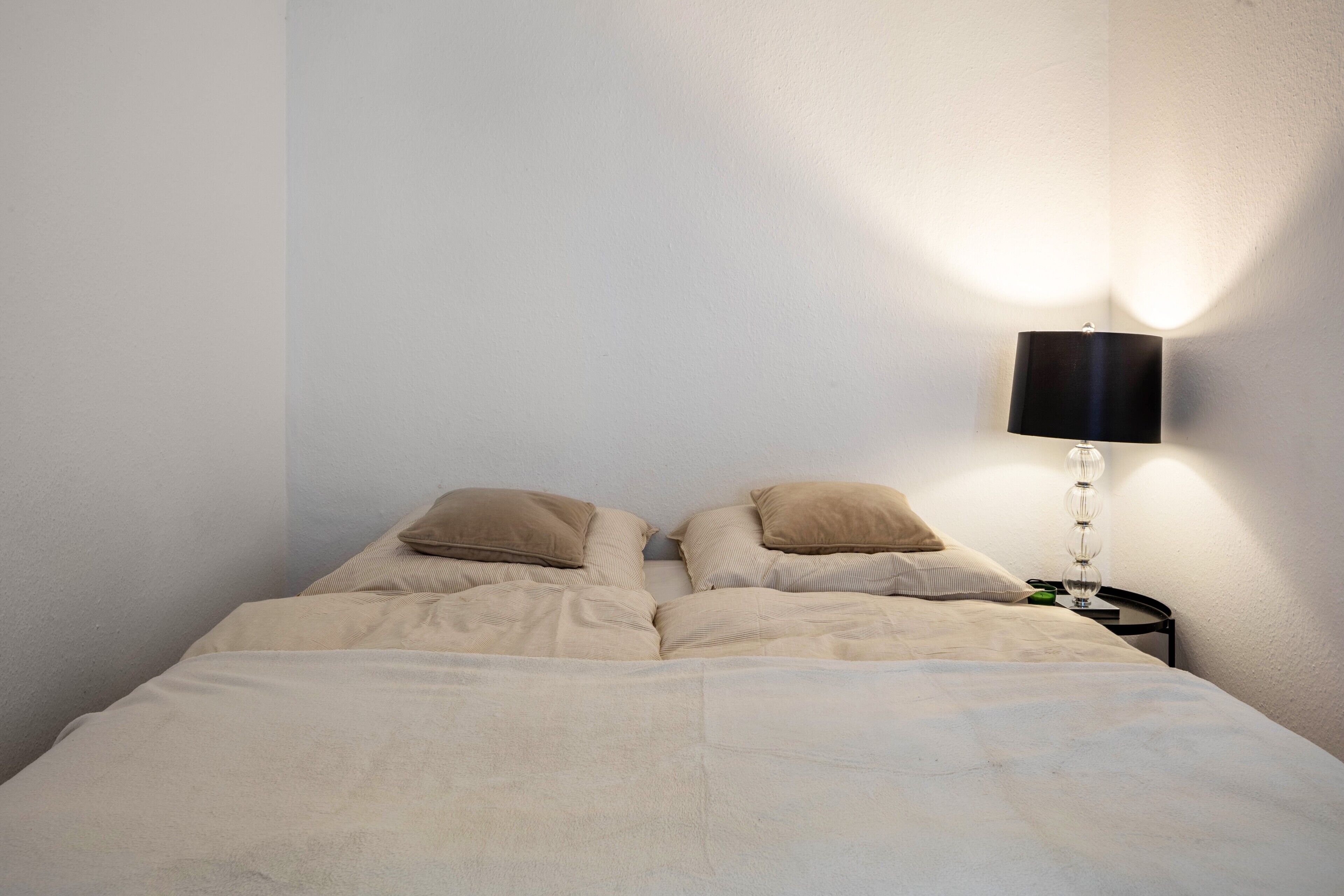 A bed with beige bedding, two pillows, and a bedside lamp against a plain wall.