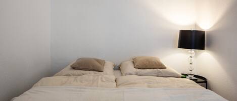 A bed with beige bedding, two pillows, and a bedside lamp against a plain wall.