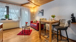 A modern living room with a red sofa, a wooden dining table, and a bar stool. There is a radiator, a small round table with a vase, and a plant. - Apartment '5' with Mountain View, Shared Terrace and Wi-Fi (Bad Rippoldsau-Schapbach)