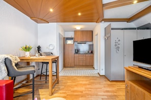 A compact kitchen with wooden cabinets, a microwave, and a stove. There is a dining area with a table and chairs, and a TV on a wooden stand.