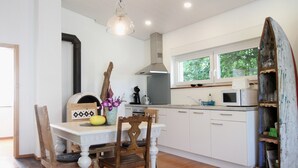 Une cuisine moderne avec des meubles blancs, une table à manger avec des chaises, un luminaire suspendu et une fenêtre donnant sur un paysage verdoyant.