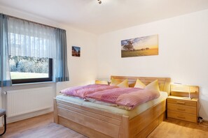 A bedroom with a wooden bed, a bedside table with a lamp, a window with curtains, and two paintings on the wall.