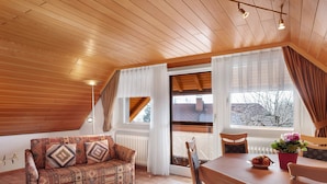 Een houten plafond, een bank met gekleurde kussens, een eettafel met stoelen en een raam met gordijnen.