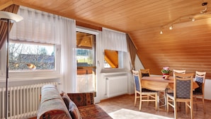 Een gezellige kamer met een houten plafond, een eettafel met stoelen, een bank en een raam met gordijnen.