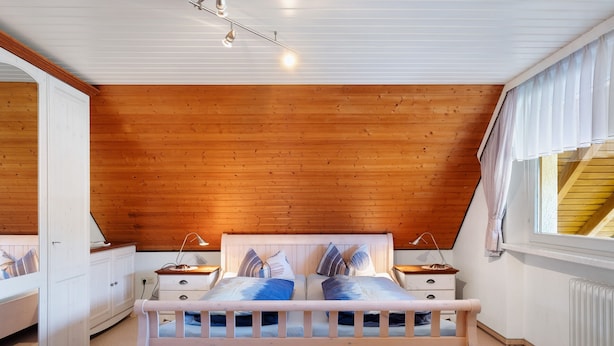 Een slaapkamer met een houten plafond, een bed met kussens, nachtkastjes, een raam met gordijnen en een radiator.