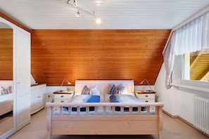 Une chambre avec un plafond en bois, un lit avec des oreillers, des tables de chevet, une fenêtre avec des rideaux et un radiateur.