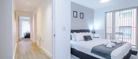 A modern bedroom with a large bed, a view through a window, and a hallway leading to another room.