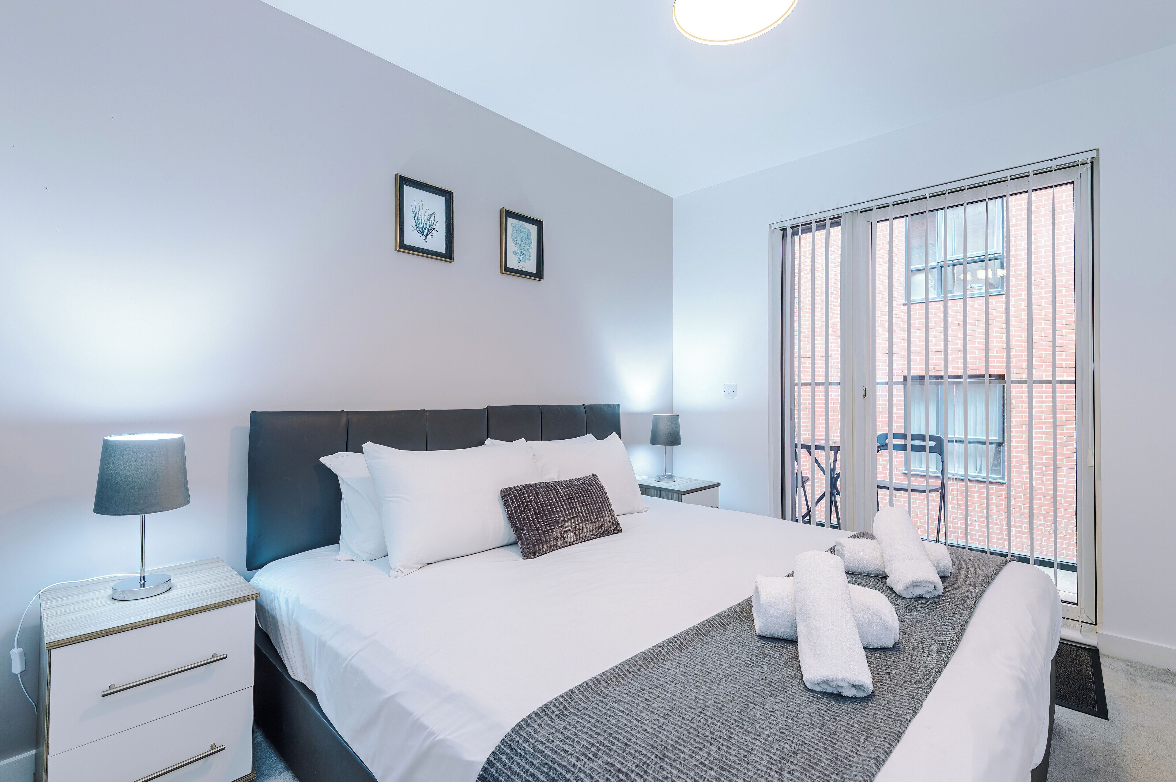 Ein modernes Hotelzimmer mit einem ordentlich bezogenen Bett, einem Nachttisch mit Lampe, einem Fenster mit Blick auf ein Backsteingebäude und zwei gerahmten Bildern an der Wand.