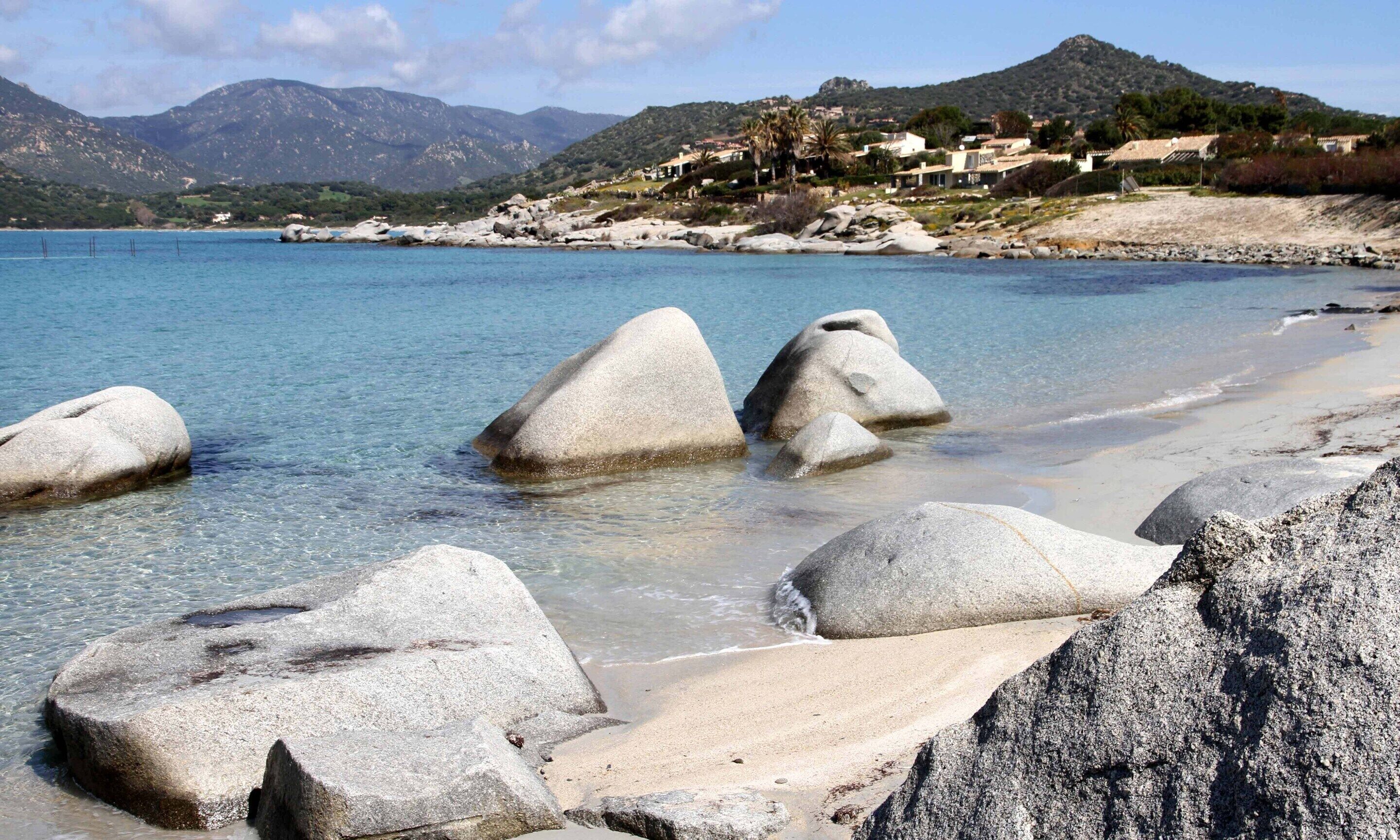 A coastal scene with large boulders in shallow water, a clear blue sea, and a mountainous backdrop with buildings.