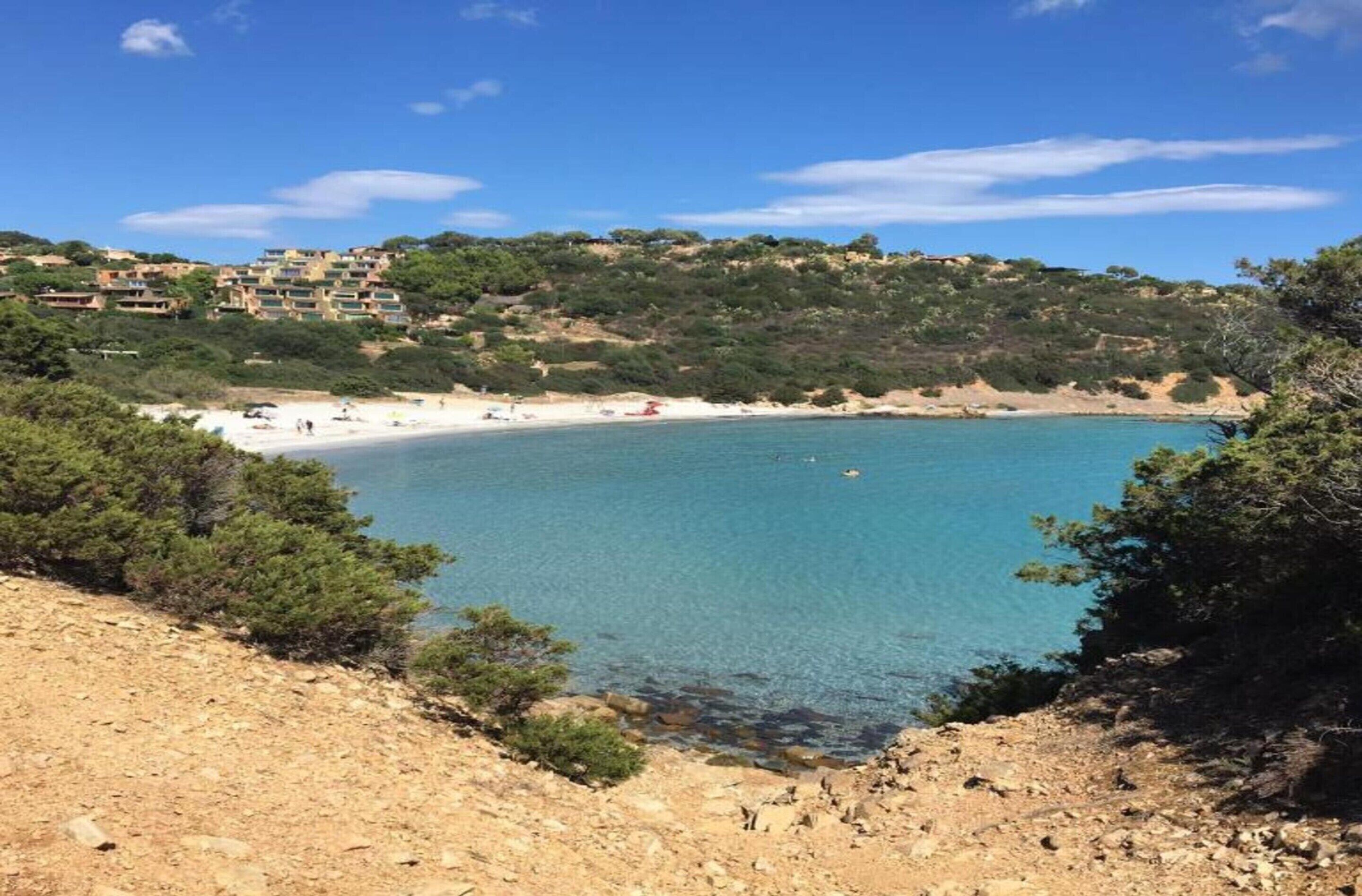 Et kystområde med en sandstrand, klart blåt vand og bygninger på en skråning.