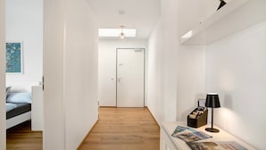 A hallway with a wooden floor, a bed with a white headboard, a door, a wall-mounted light fixture, and a magazine rack.