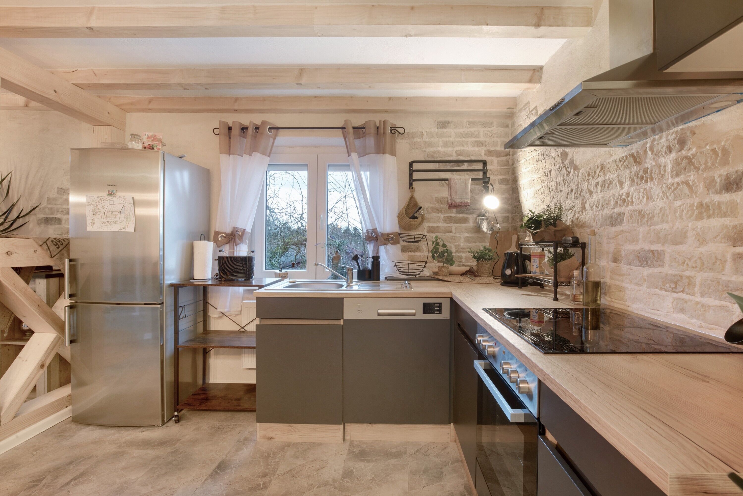 Cocina compacta con lavavajillas integrado, refrigerador de acero inoxidable, estufa y ventana con cortinas.