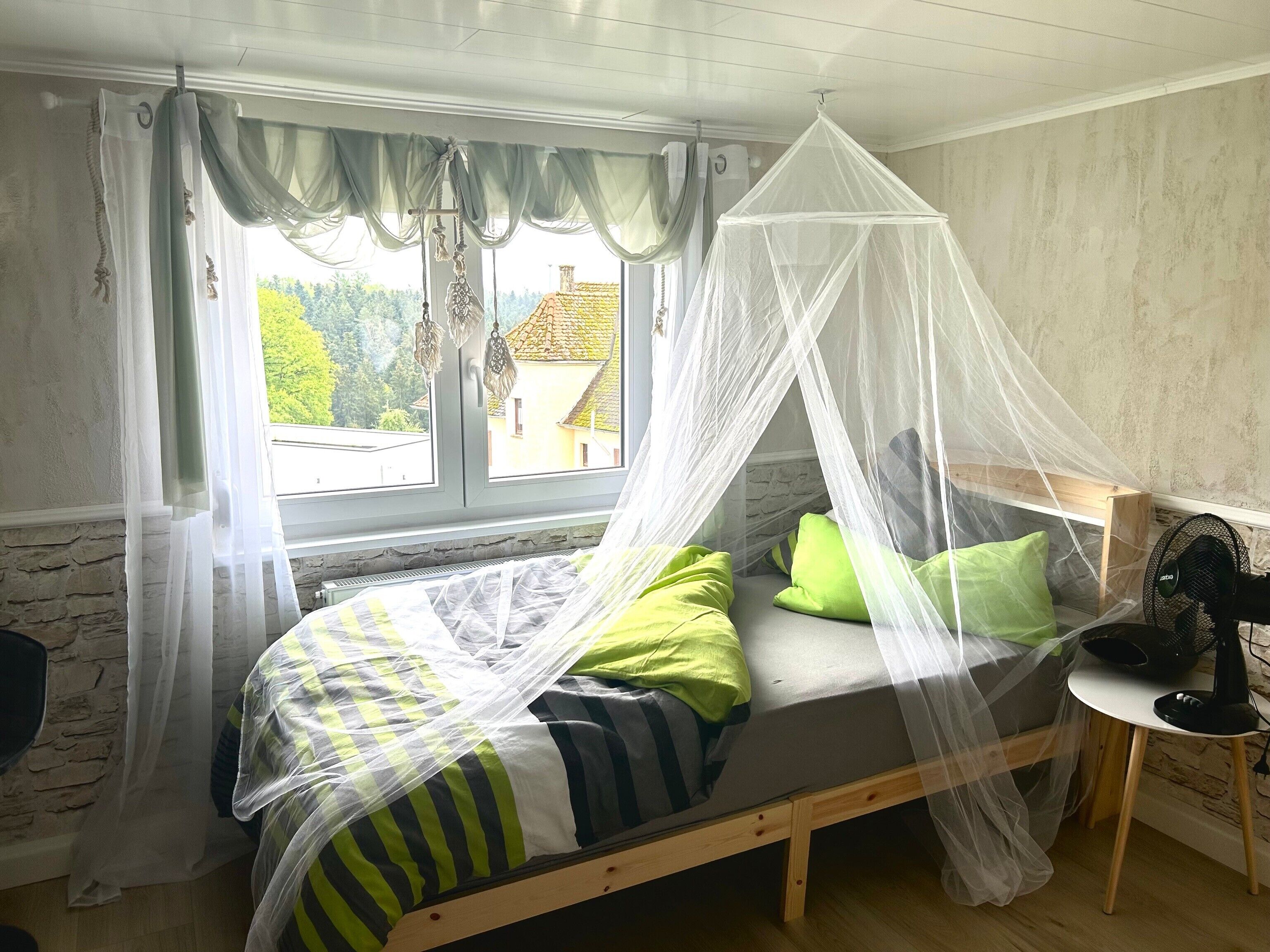 Ein Zimmer mit einem Bett, einem Nachttisch mit Ventilator, einem Fenster mit Vorhängen und Blick auf Gebäude.