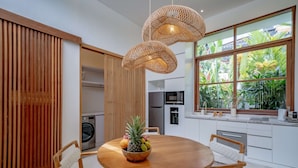 Eine moderne Küche mit einem runden Holztisch, zwei Weidenkorb-Pendelleuchten und Blick auf üppiges Grün durch große Fenster.