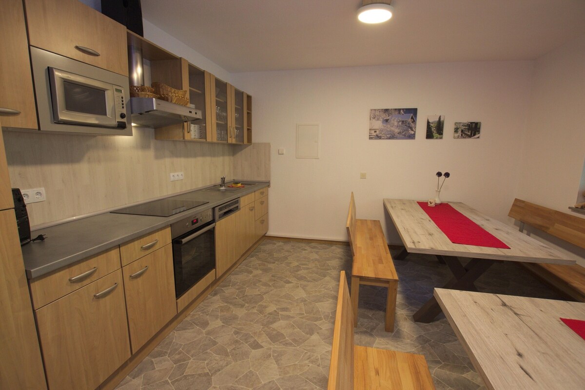 A kitchen with a microwave, oven, and wooden cabinets. There is a dining area with a table and benches.