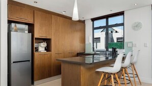 A modern kitchen with wooden cabinets, a stainless steel refrigerator, a glass island with white chairs, and a large window offering a view of a building outside.