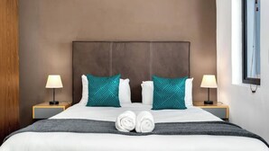 A neatly made bed with white linens and teal pillows, two bedside lamps, and a pair of rolled towels on the bed.