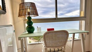 A room with a view of the sea, a table with a lamp, a chair, and a radiator.