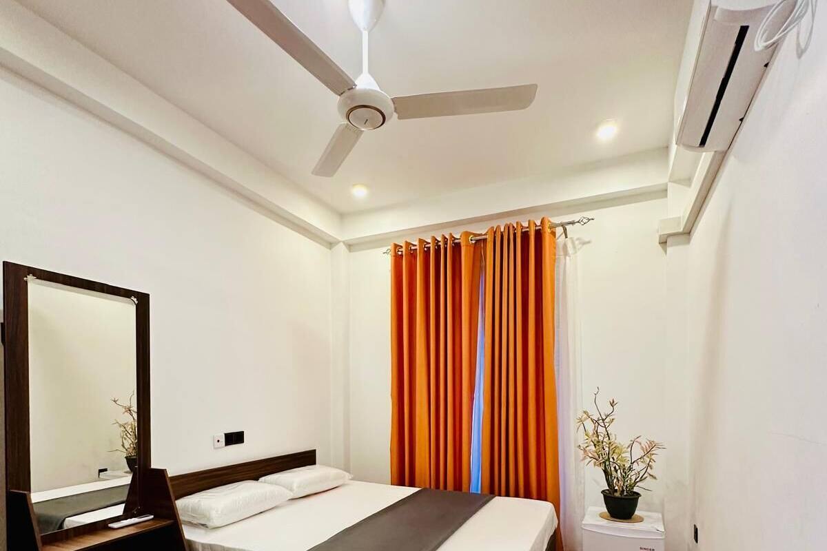 A hotel room with a bed, a ceiling fan, an air conditioner, a mirror, and a small table with a plant.