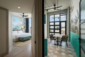 A hotel room with a bed, a small seating area, a dining table with chairs, a ceiling fan, and a large window with a city view. - Fifth Avenue Hotel (George Town)