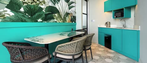 Una cocina moderna con armarios azul verdoso, microondas y cafetera. Hay una mesa de comedor con sillas y un mural con motivos vegetales en la pared.