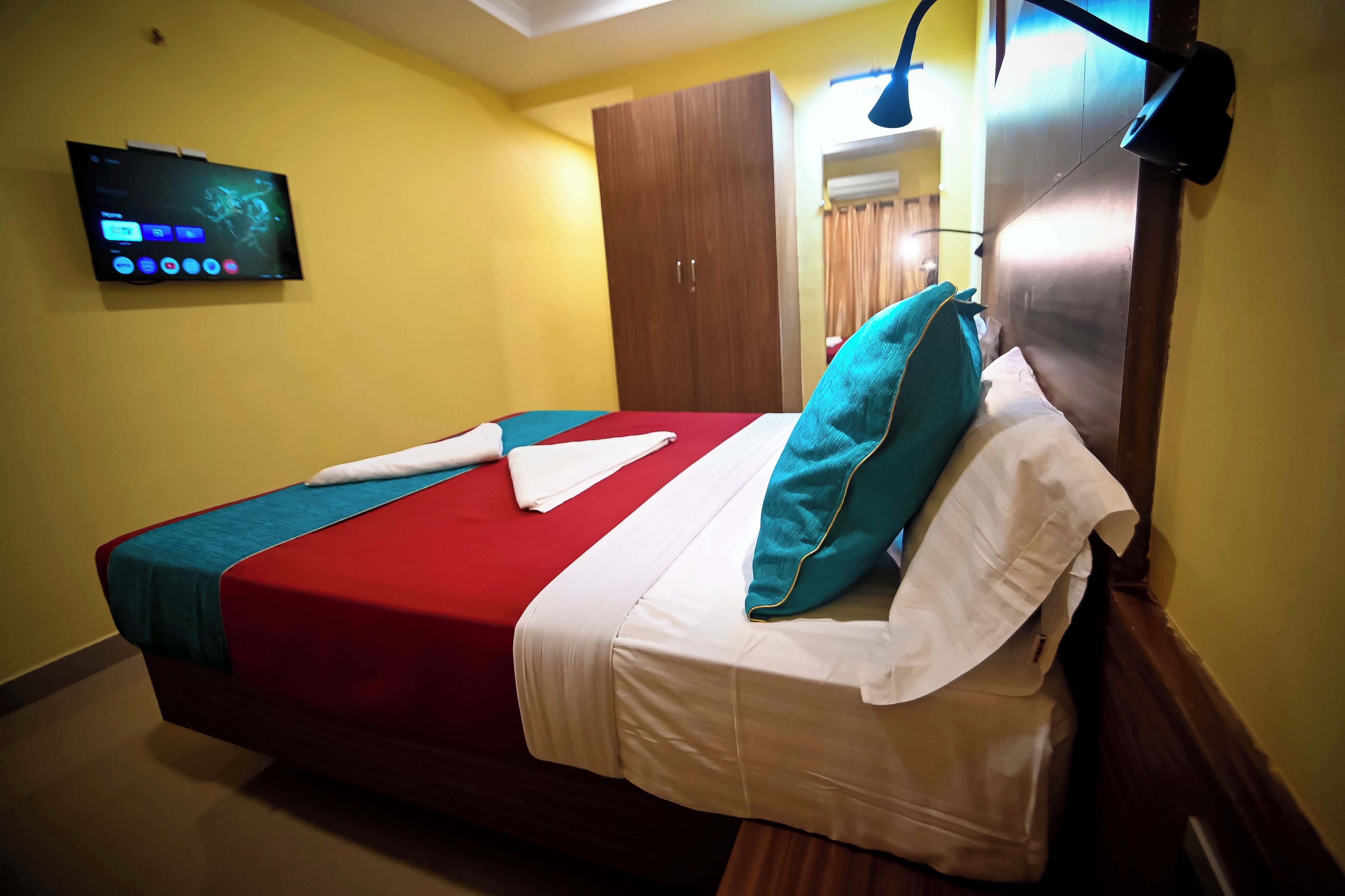 A neatly made bed with a red and white bedspread, teal pillows, and a white pillow. A flat-screen TV is mounted on the wall above the bed.