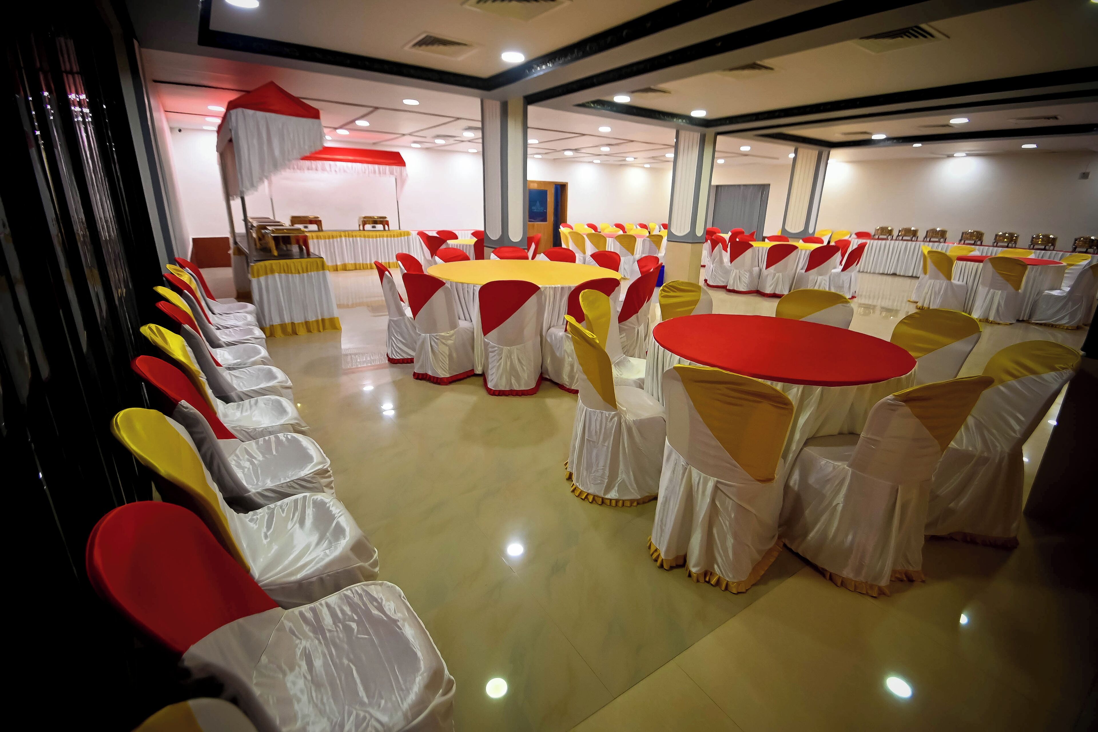 A banquet hall with round tables and chairs, some covered with white and yellow tablecloths, and red and yellow chair covers.