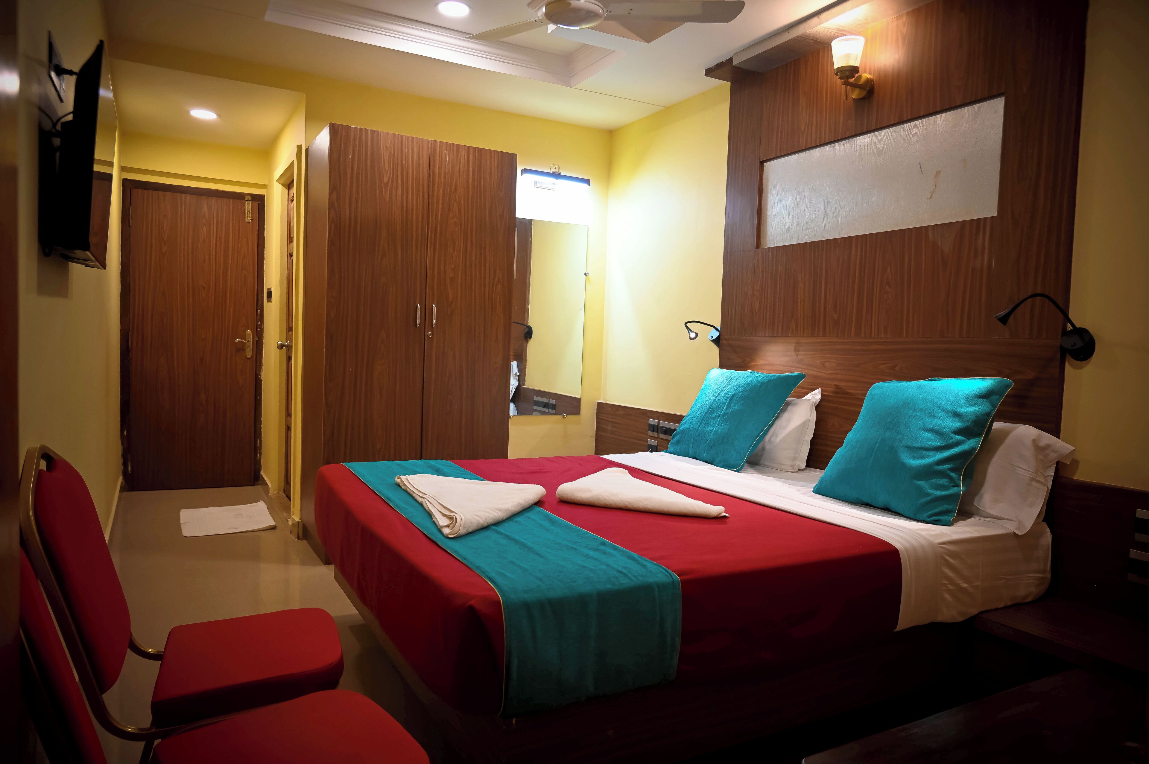 Een hotelkamer met twee bedden, rood en turquoise beddengoed, een houten hoofdbord, een flatscreen televisie en een plafondventilator.